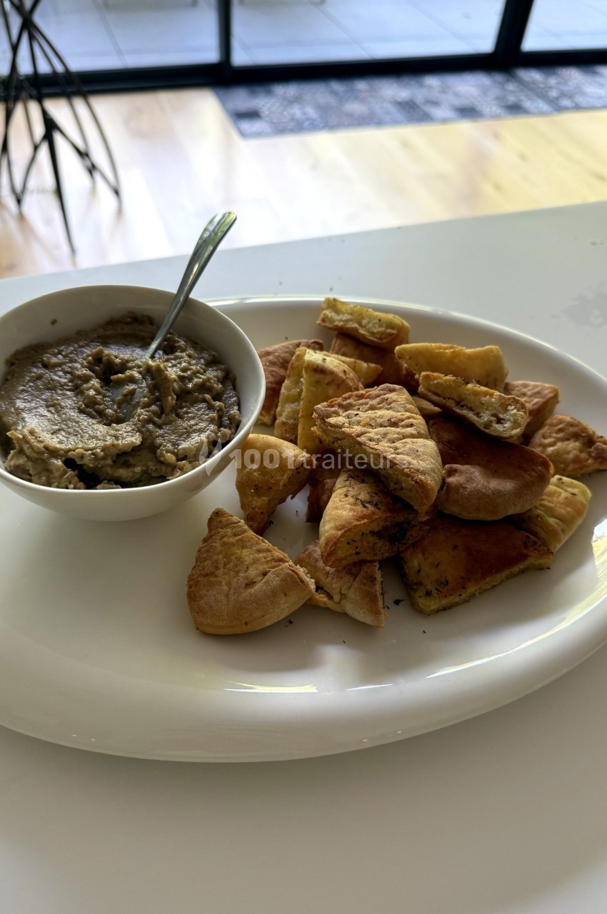 Assiette de pita grillée accompagnée d'un bol de houmous, posée sur une table blanche près d'une baie vitrée.