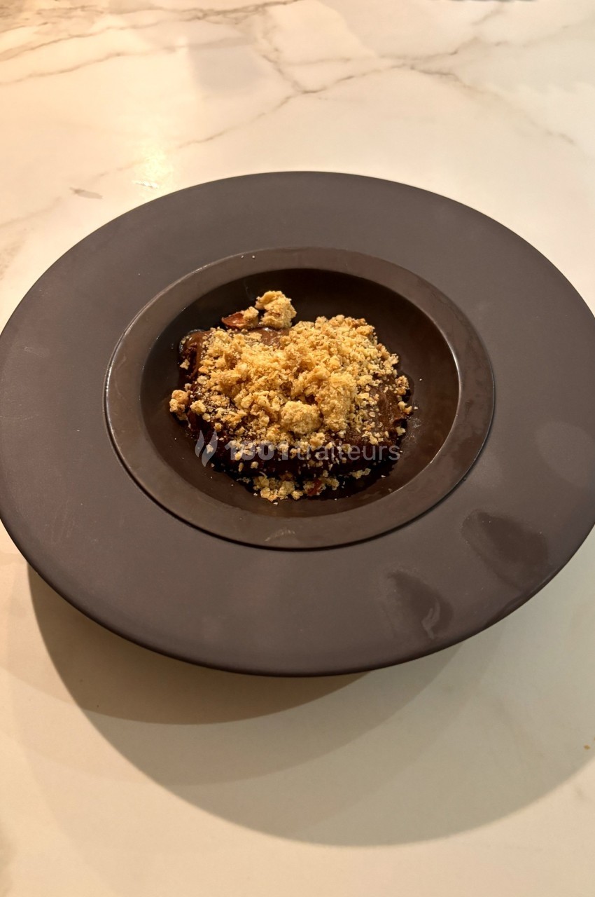 Assiette noire contenant un dessert recouvert de crumble, posée sur une table en marbre clair.