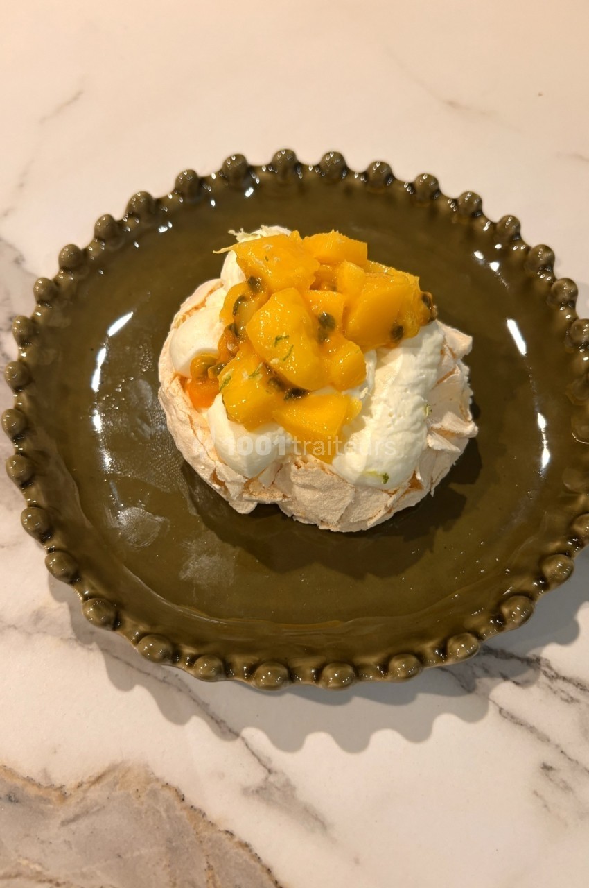 Pavlova garnie de crème fouettée, morceaux de mangue et fruits de la passion, servie sur une assiette verte.