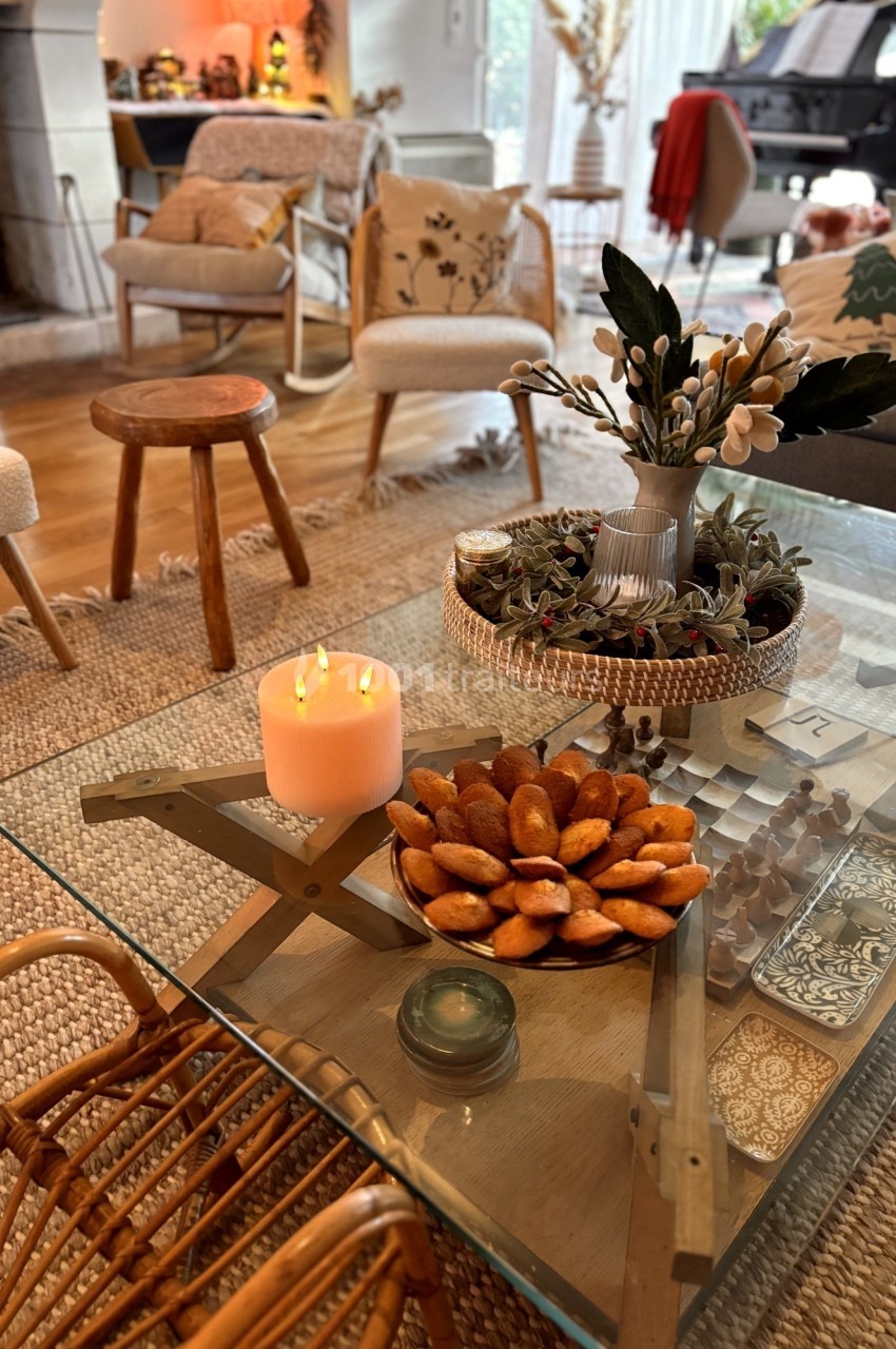 Table basse en verre décorée d'une bougie allumée, d'un plat de madeleines et d'un vase avec des branches, dans un salon…