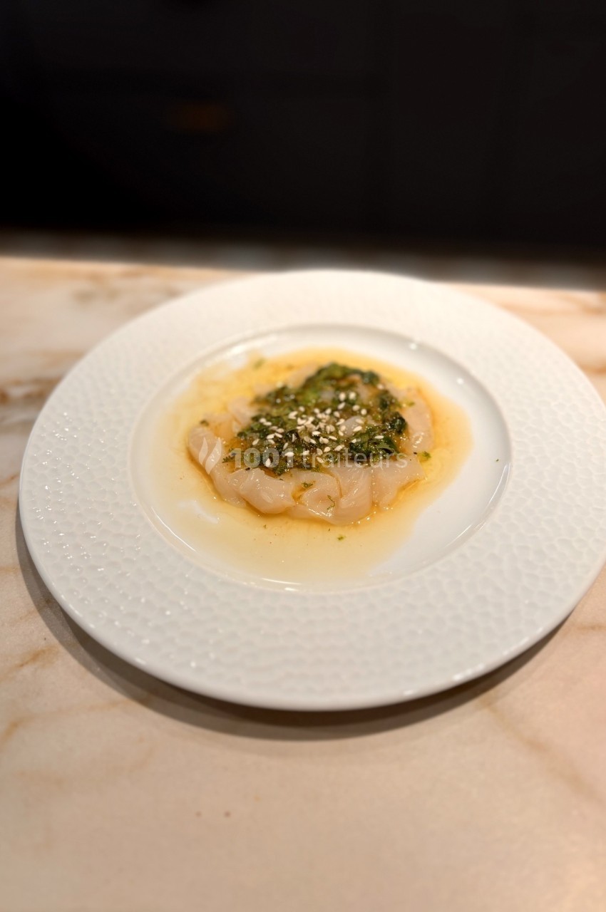 Assiette de carpaccio de poisson blanc avec sauce verte et graines de sésame, servie sur une table en marbre.