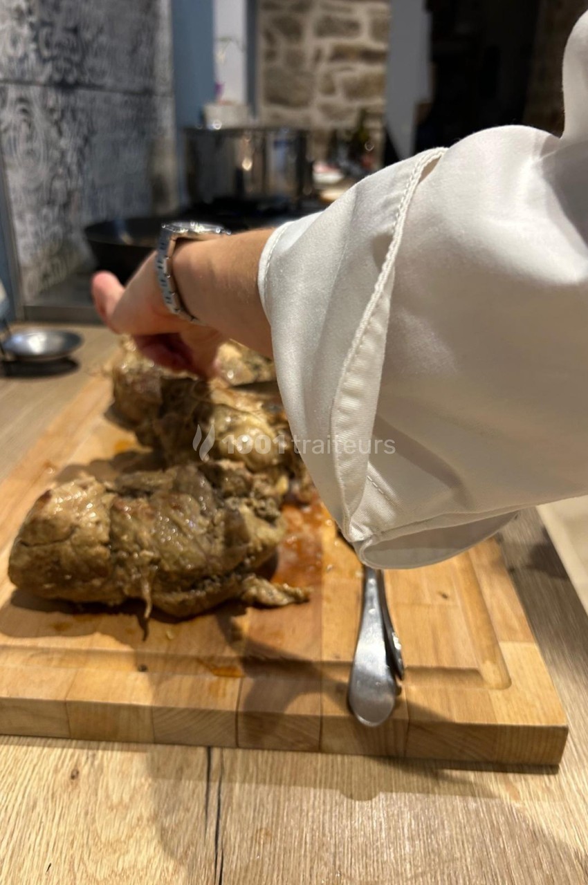 Une main en chemise blanche prépare des morceaux de viande cuite sur une planche en bois dans une cuisine.