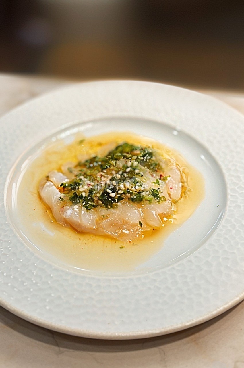 Carpaccio de poisson blanc finement tranché, assaisonné d'herbes, d'épices et d'huile, servi sur une assiette blanche.