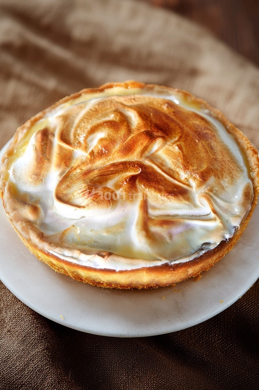 Tartelette au citron meringuée dorée, posée sur une assiette blanche avec un fond en tissu brun.