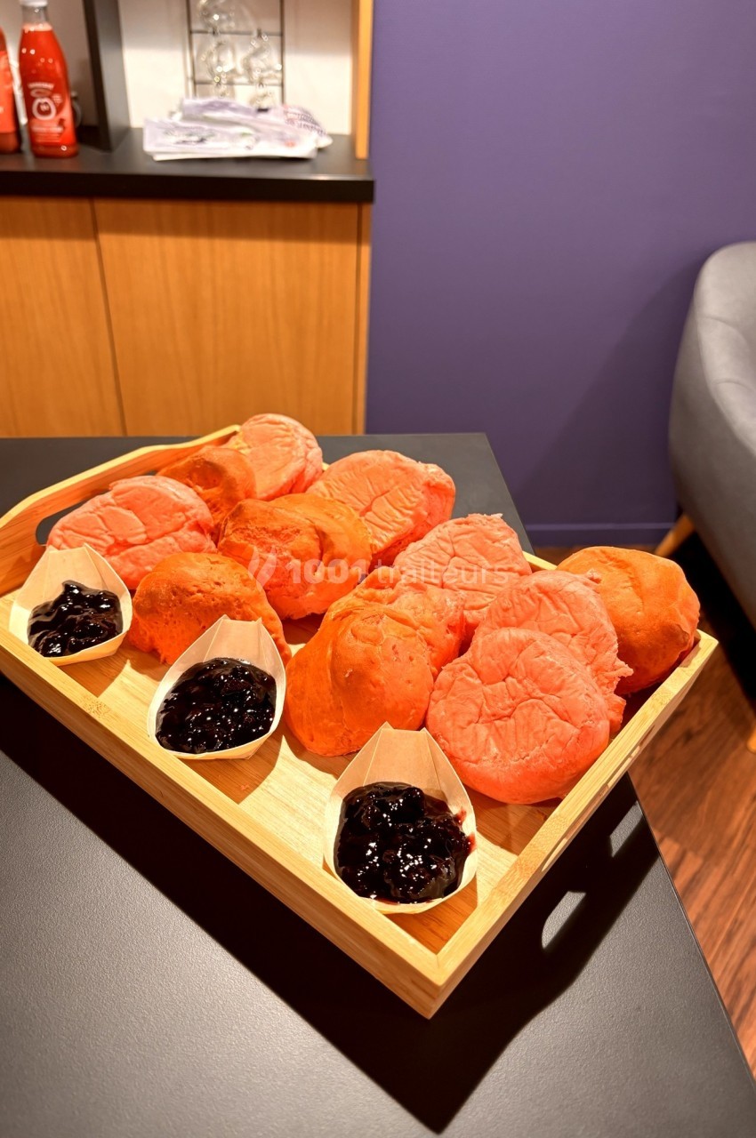 Plateau en bois contenant des petits pains roses accompagnés de confitures servies dans des coupelles en papier.