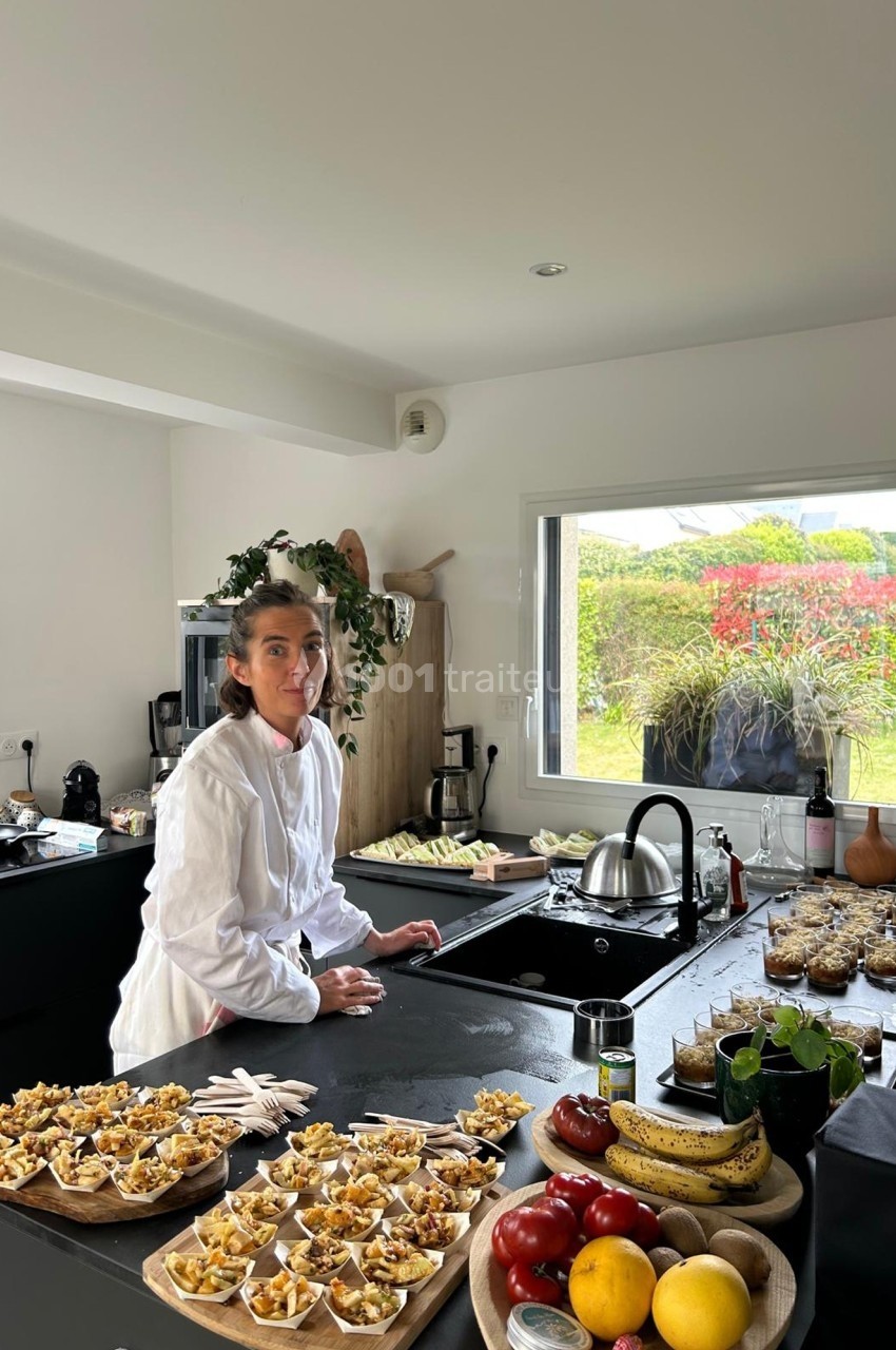 Une femme en blouse blanche prépare des plats dans une cuisine moderne avec vue sur un jardin fleuri.