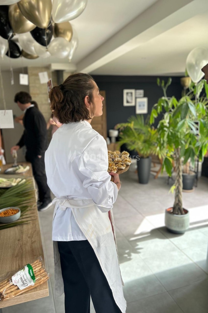 Une personne en blouse blanche tient un plateau de nourriture dans une pièce décorée avec des ballons et des plantes.