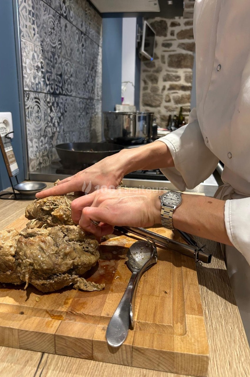 Une personne découpe un morceau de viande cuite sur une planche en bois dans une cuisine au mur en pierre.