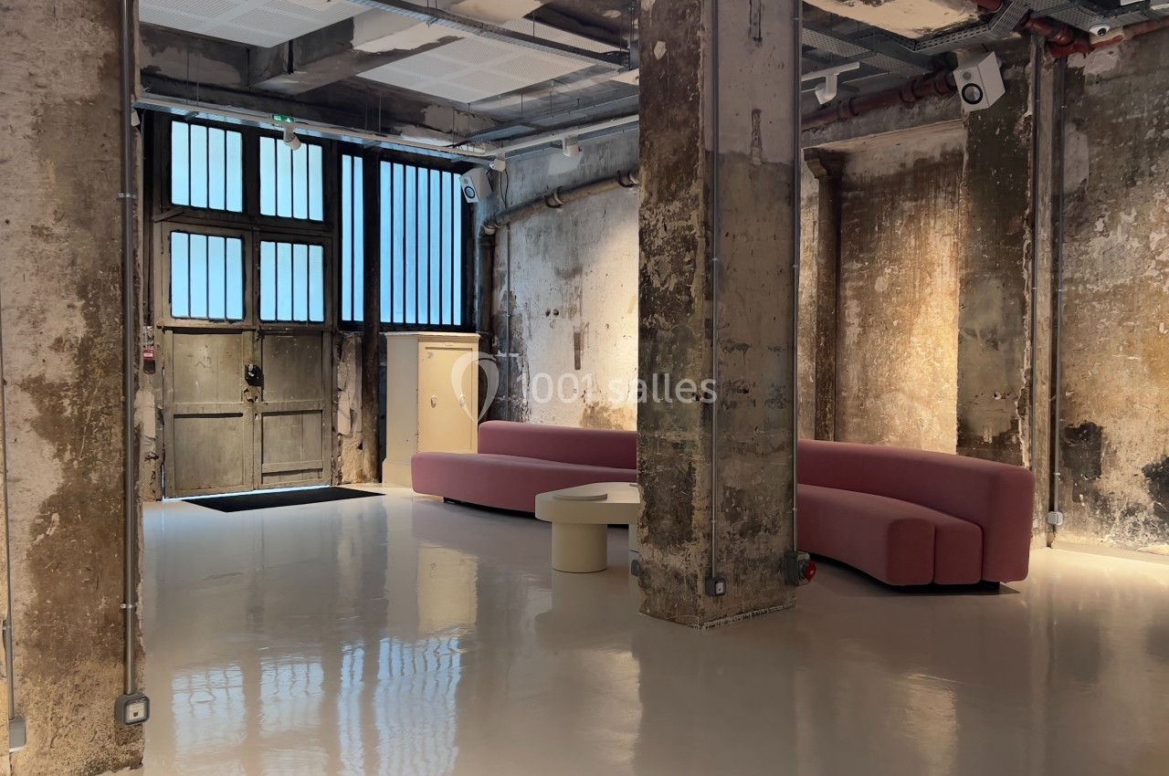 Espace intérieur industriel avec murs bruts, colonnes en béton, canapé rose et sol brillant.