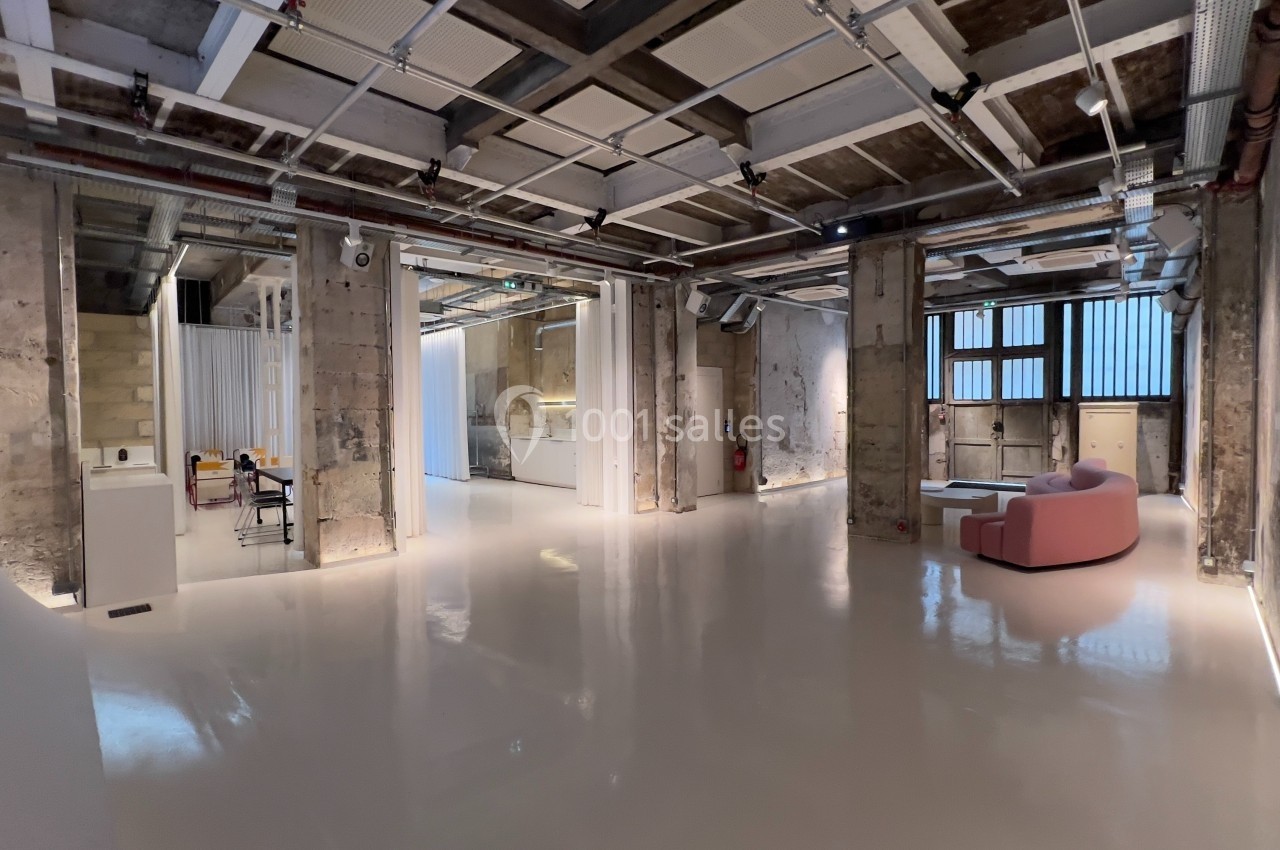 Espace intérieur industriel rénové avec murs en béton brut, sol brillant, mobilier moderne et grandes fenêtres.