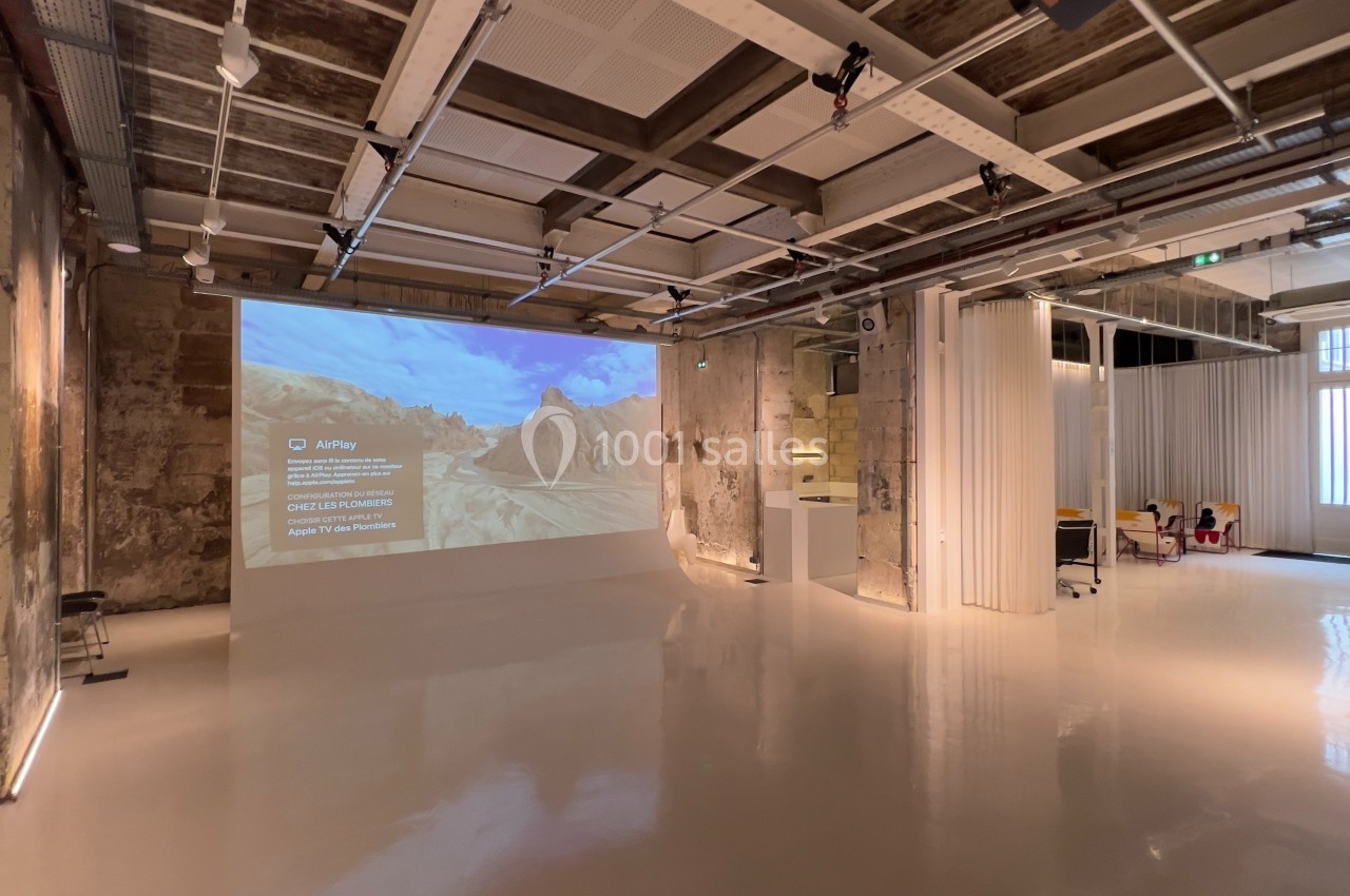 Espace intérieur moderne avec murs en béton brut, sol brillant, écran de projection et mobilier minimaliste.
