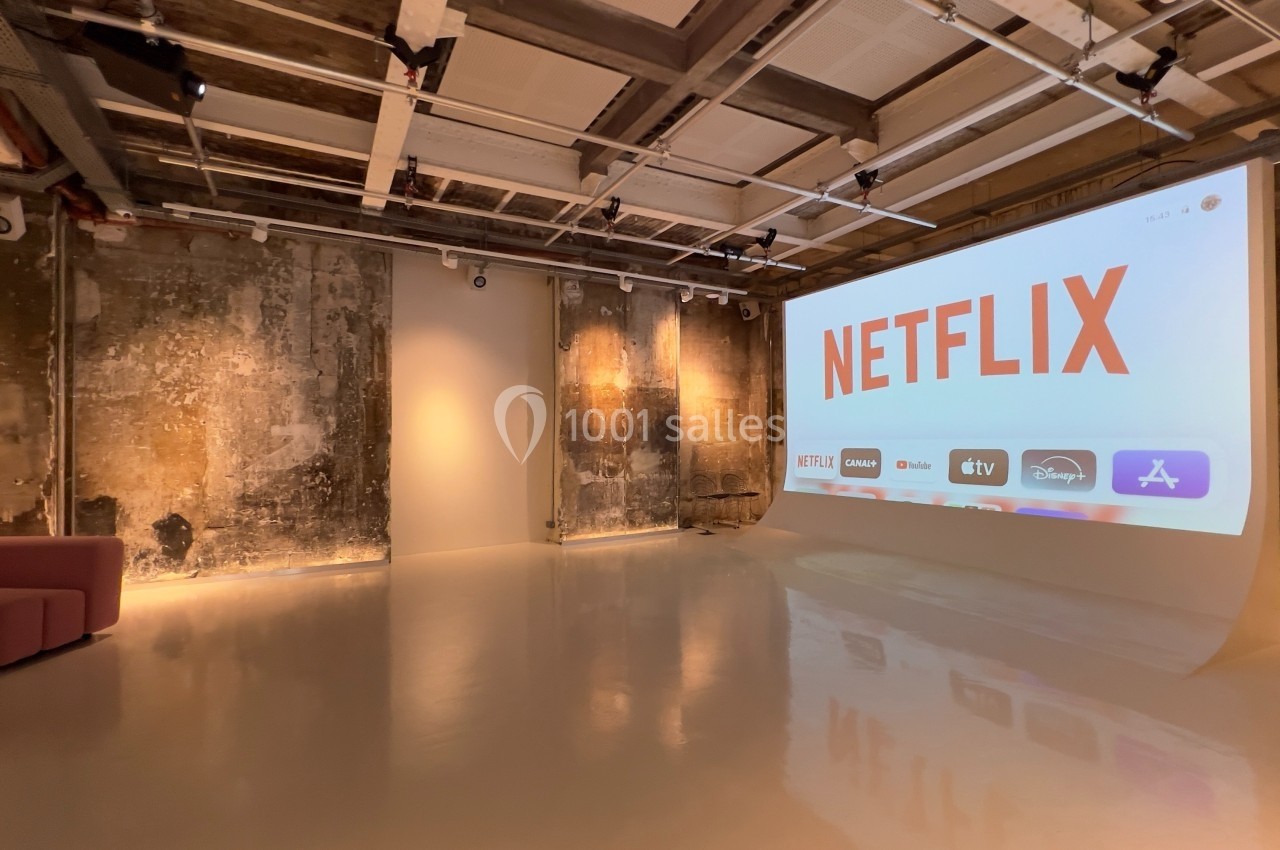 Salle moderne avec un grand écran affichant le logo Netflix et des icônes de plateformes de streaming, murs bruts en arrière…