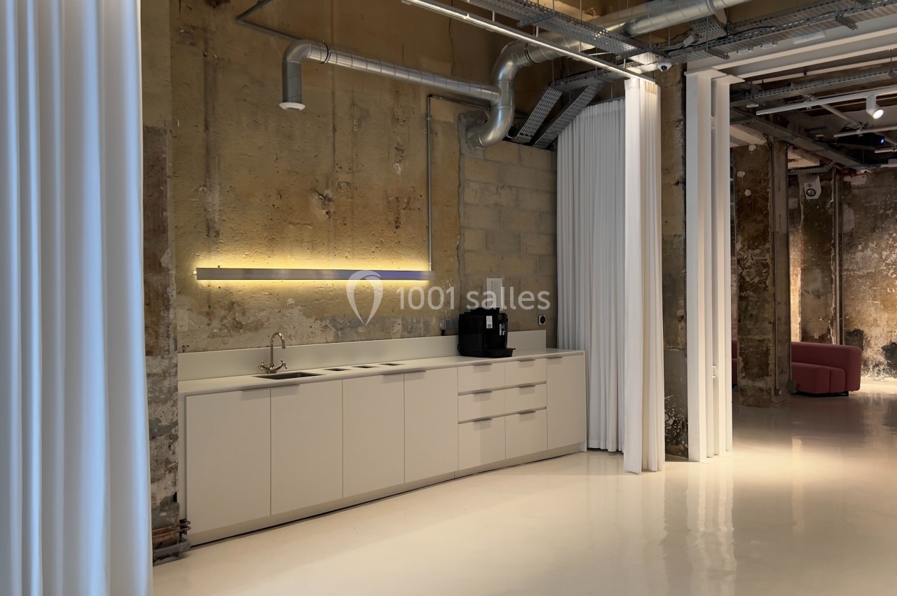 Cuisine moderne avec placards blancs, évier, machine à café et éclairage mural dans un espace industriel avec murs bruts.