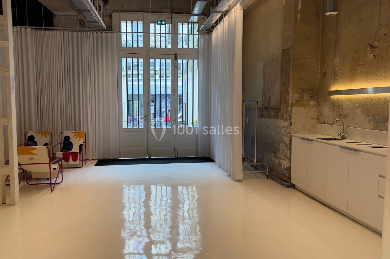 Espace intérieur lumineux avec sol blanc, murs bruts, kitchenette moderne et grandes portes vitrées donnant sur l'extérieur.