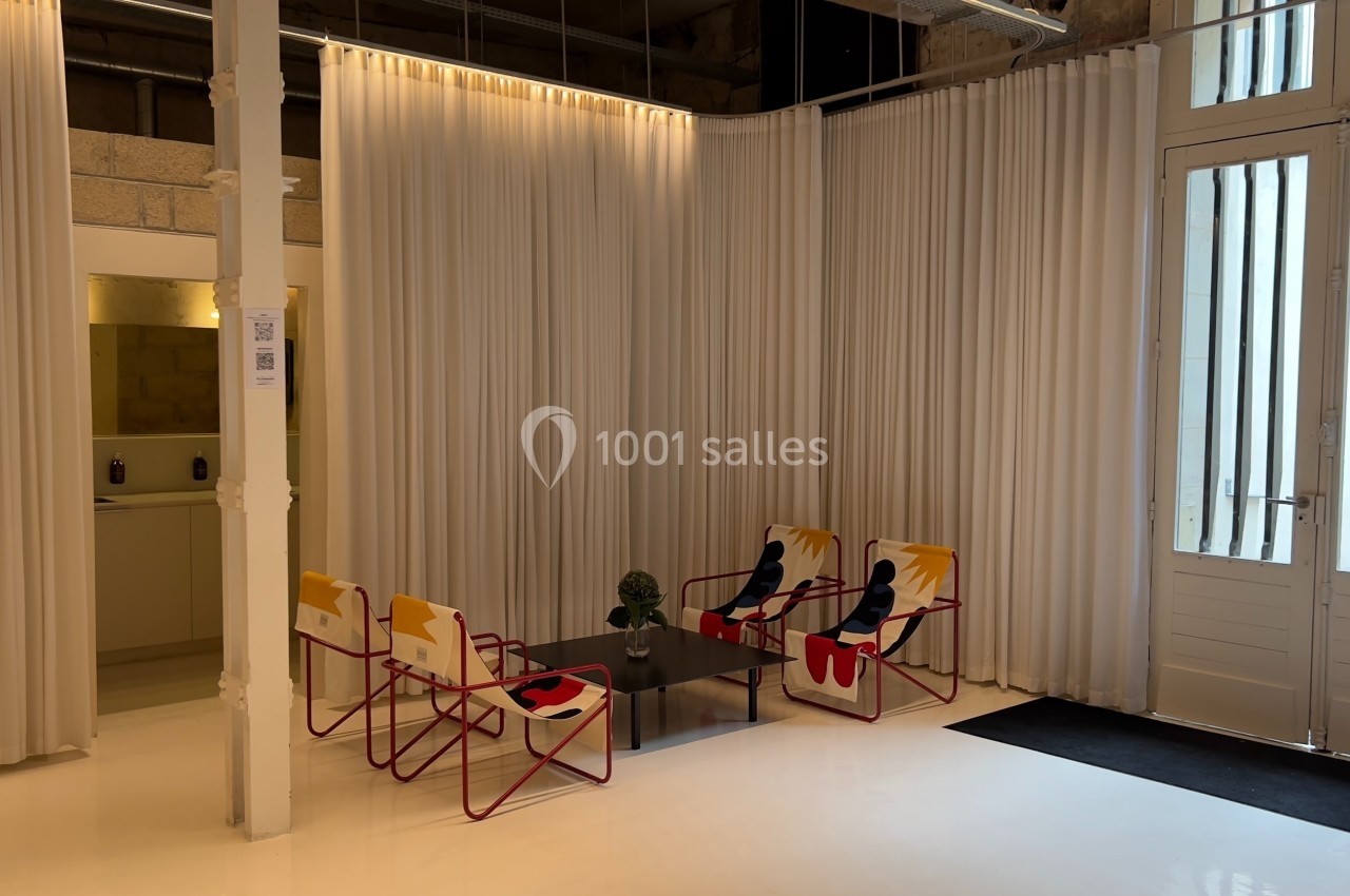 Espace lumineux avec des chaises colorées autour d'une table basse, entouré de rideaux blancs et d'une porte vitrée.