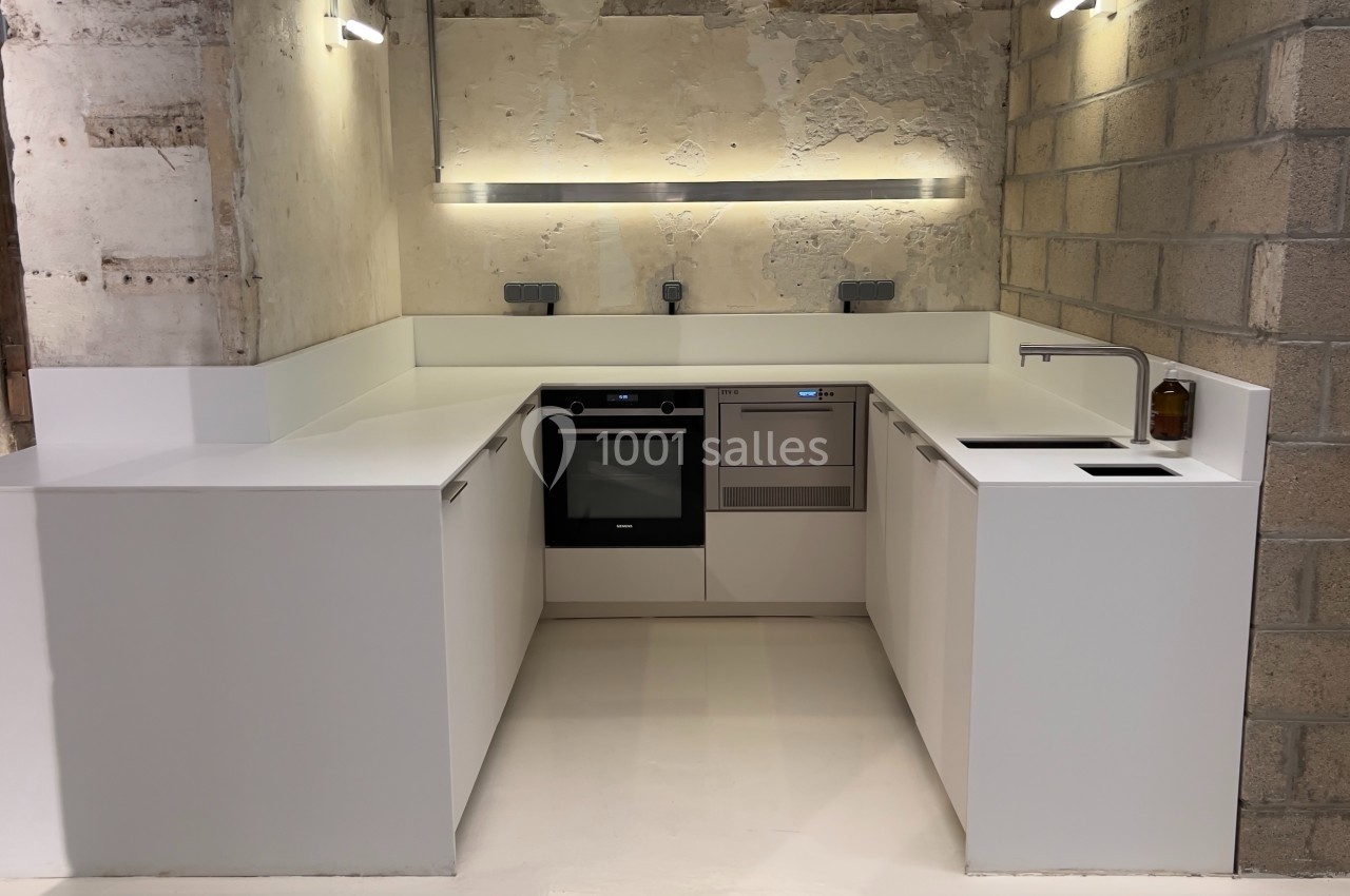 Cuisine moderne en U avec meubles blancs, plan de travail épuré, mur en béton brut et éclairage intégré.