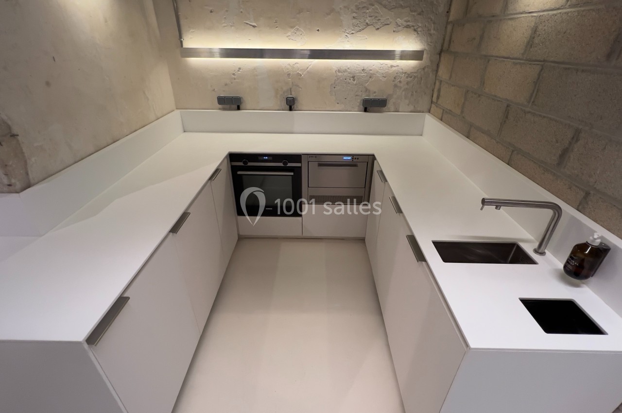 Cuisine moderne en angle avec plan de travail blanc, évier intégré, plaques de cuisson et murs en béton brut.