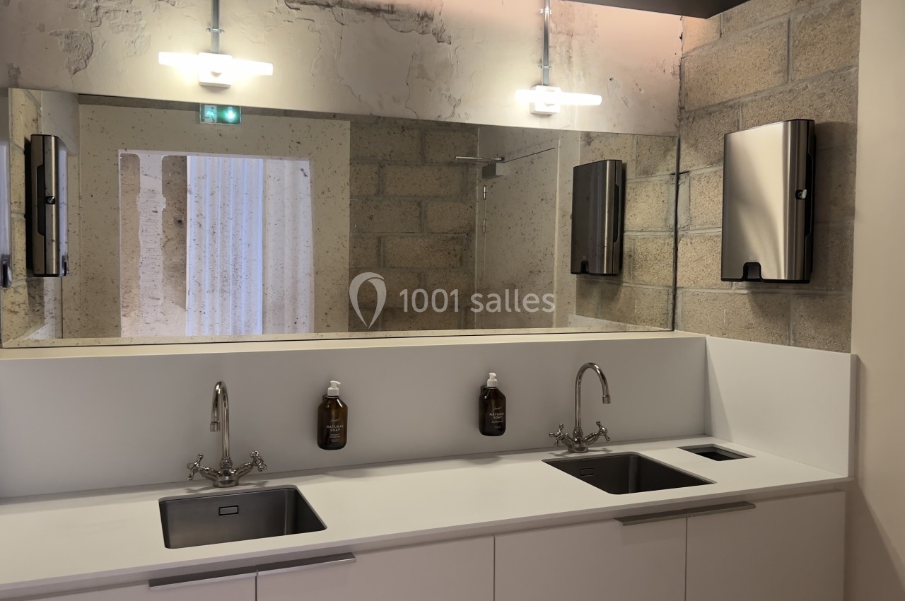 Deux éviers en inox avec distributeurs de savon, miroir et murs en béton dans une salle de bain moderne.