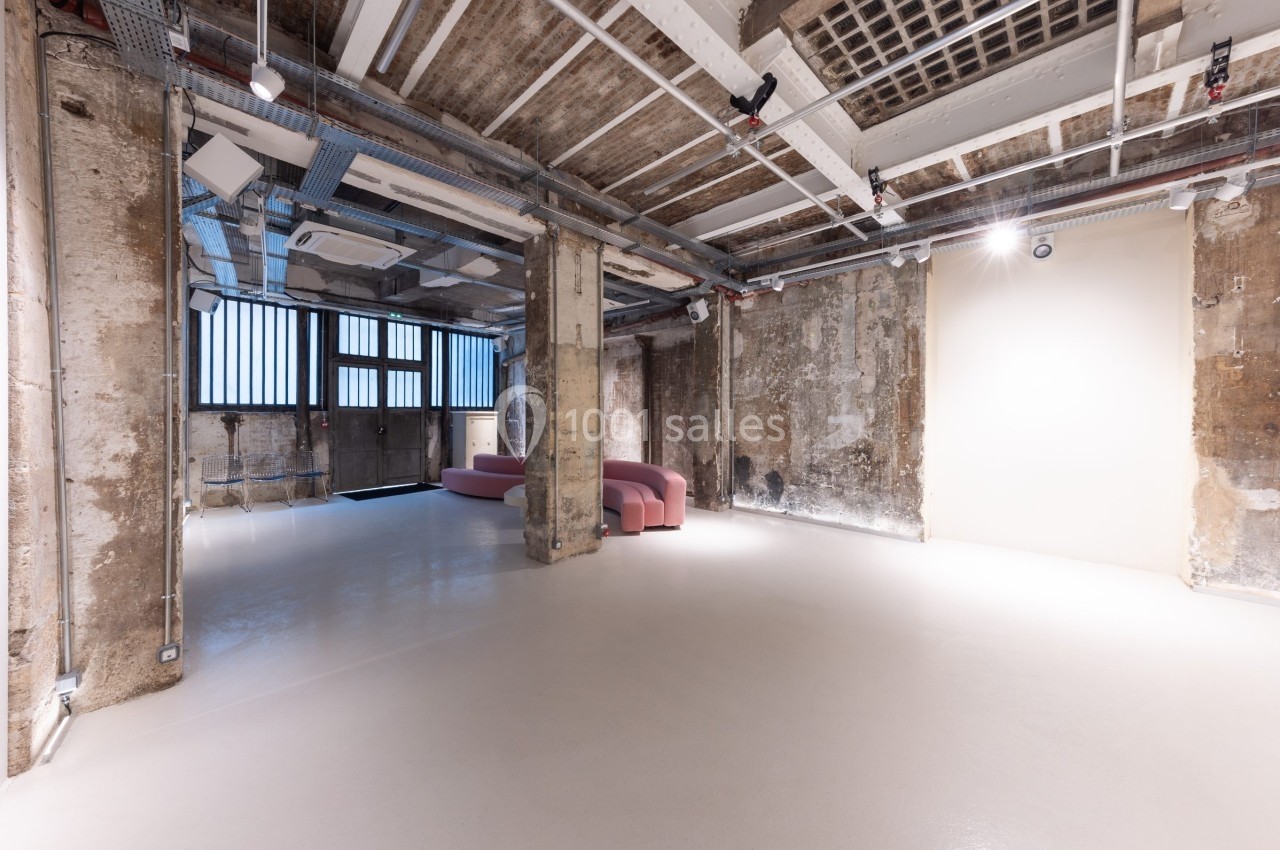 Espace intérieur industriel avec murs en béton brut, sol clair, canapé rose et grandes fenêtres à barreaux.