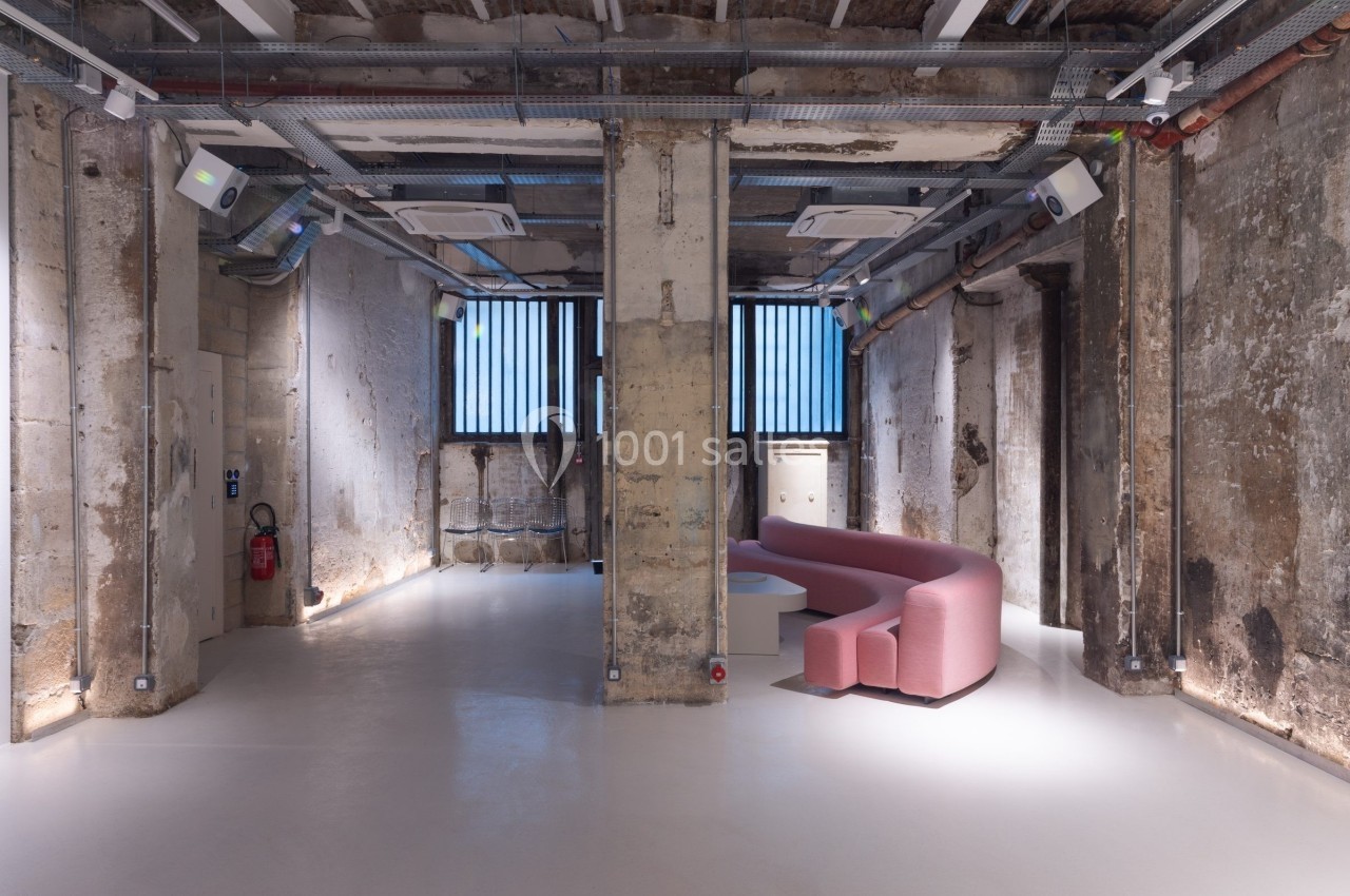 Espace intérieur industriel rénové avec murs en béton brut, fenêtres à barreaux et canapé rose incurvé.