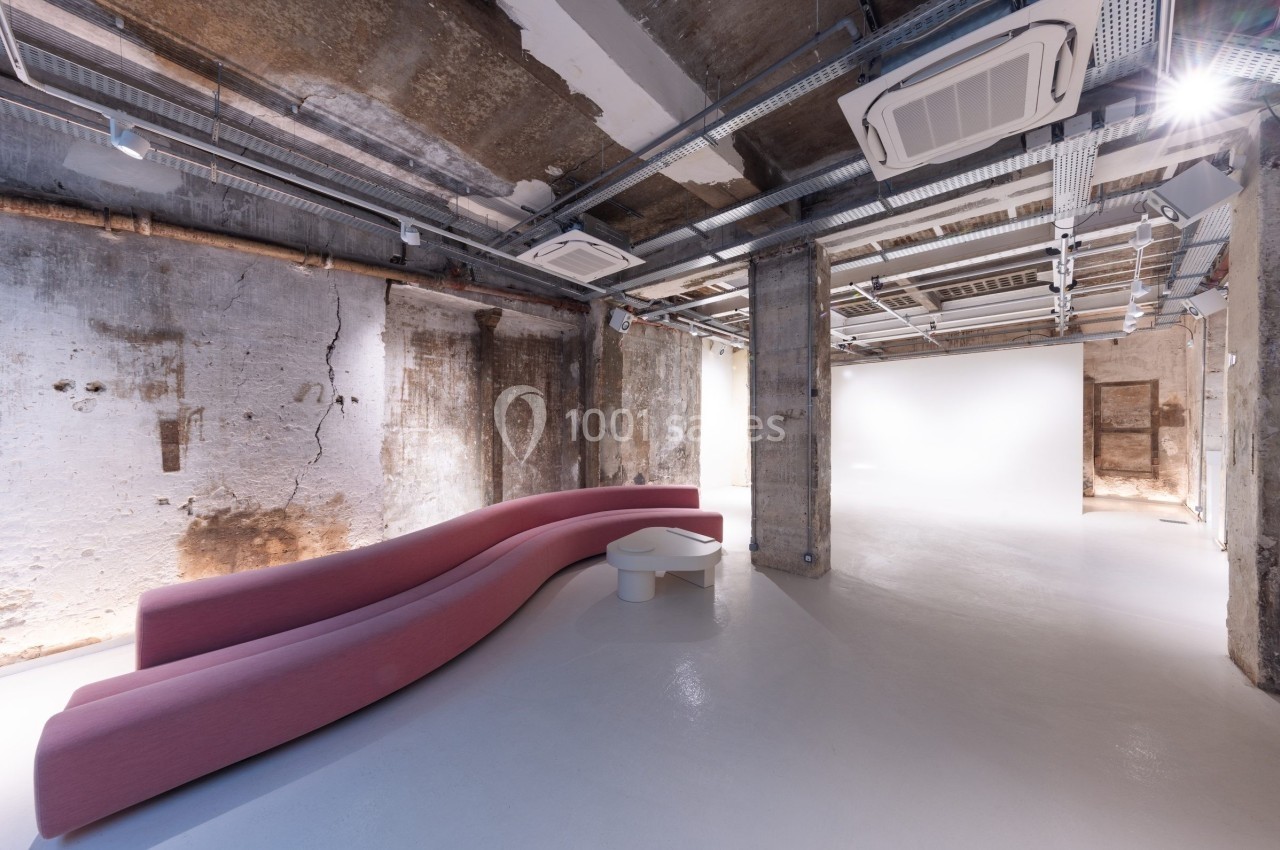 Espace intérieur industriel avec murs bruts, plafond apparent et canapé rose incurvé sur un sol blanc.