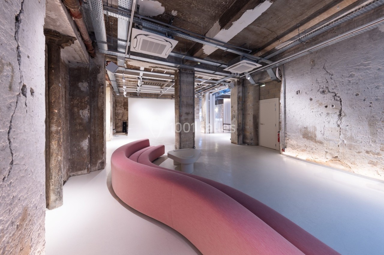 Espace intérieur industriel avec murs en béton brut, plafond technique apparent et canapé rose incurvé.
