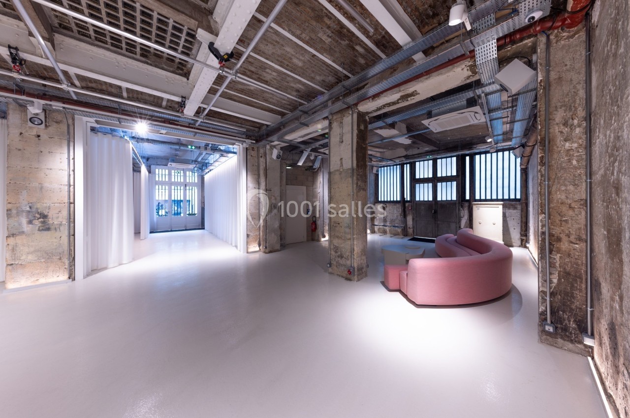 Espace intérieur industriel avec murs en béton brut, plafond exposé et canapé rose dans un coin lumineux.