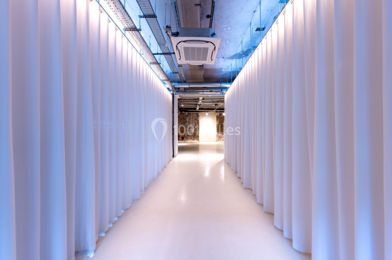 Couloir lumineux avec sol blanc, rideaux blancs de chaque côté et plafond industriel exposant des conduits.