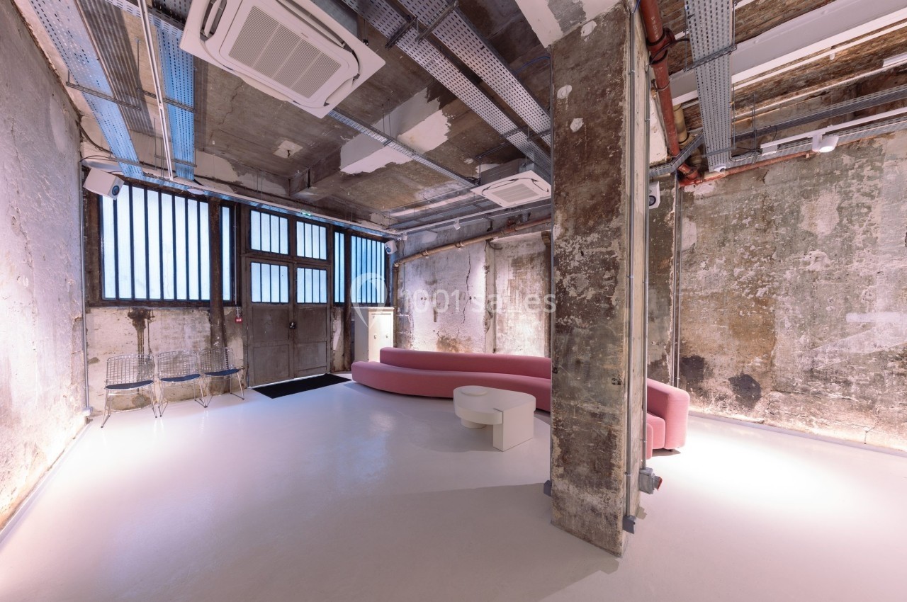 Espace intérieur industriel rénové avec murs bruts, canapé rose, chaises transparentes et lumière naturelle.