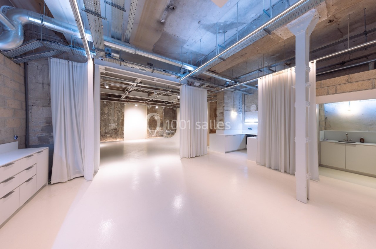 Espace intérieur moderne avec sol clair, murs en béton brut, rideaux blancs et éclairage industriel.