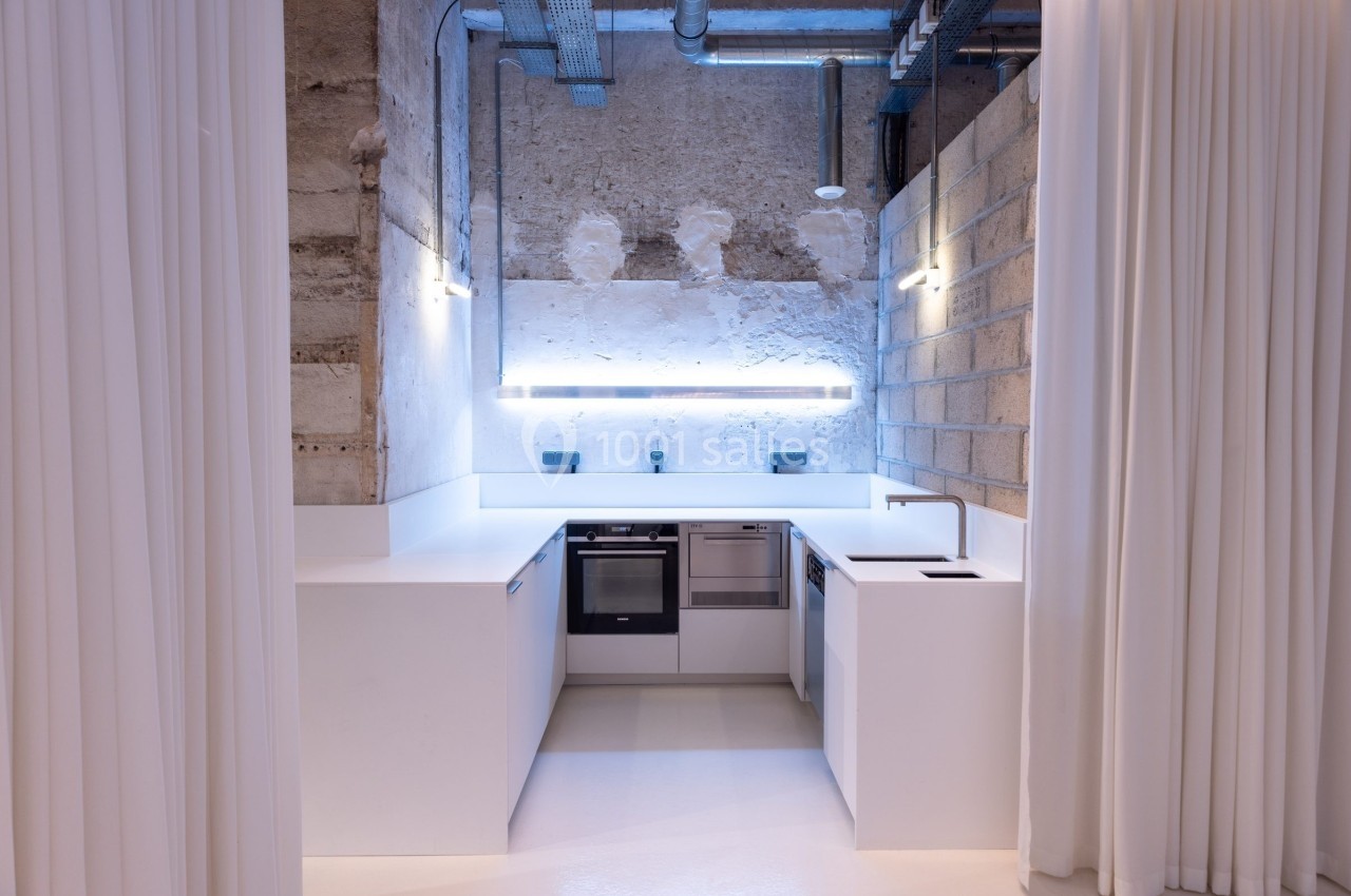 Cuisine moderne avec murs en béton brut, éclairage LED, plan de travail blanc, et appareils encastrés.