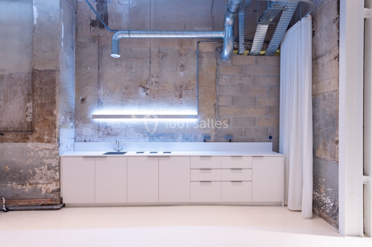 Cuisine moderne minimaliste avec meubles blancs, éclairage LED et murs en béton brut dans un espace industriel.