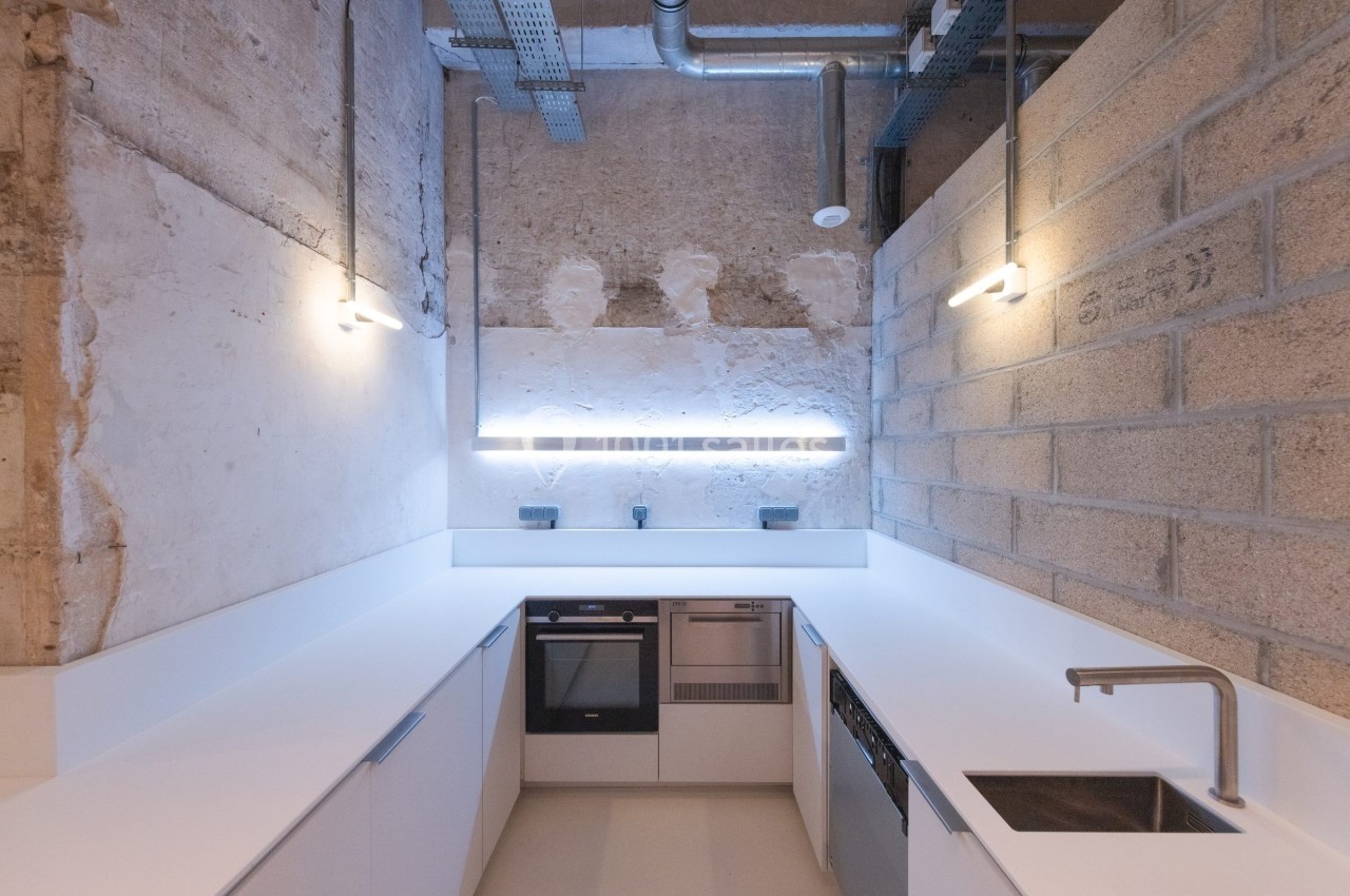 Cuisine moderne avec murs en béton brut, éclairage minimaliste, plan de travail blanc et électroménagers intégrés.