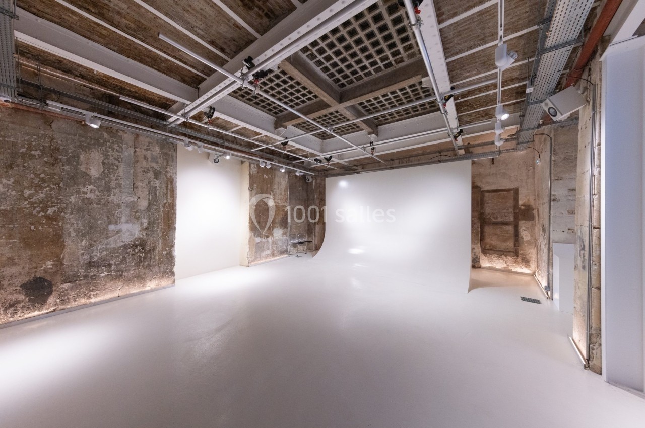 Studio photo avec murs bruts, sol blanc, éclairage au plafond et fond blanc pour prises de vue.