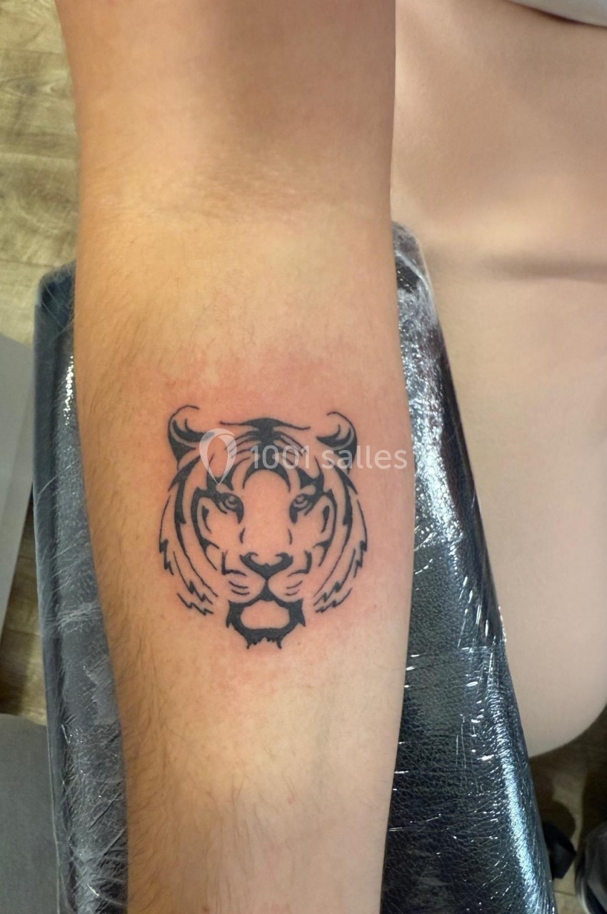 Tatouage noir d'une tête de tigre sur l'avant-bras d'une personne, posé sur un appui recouvert de film plastique.