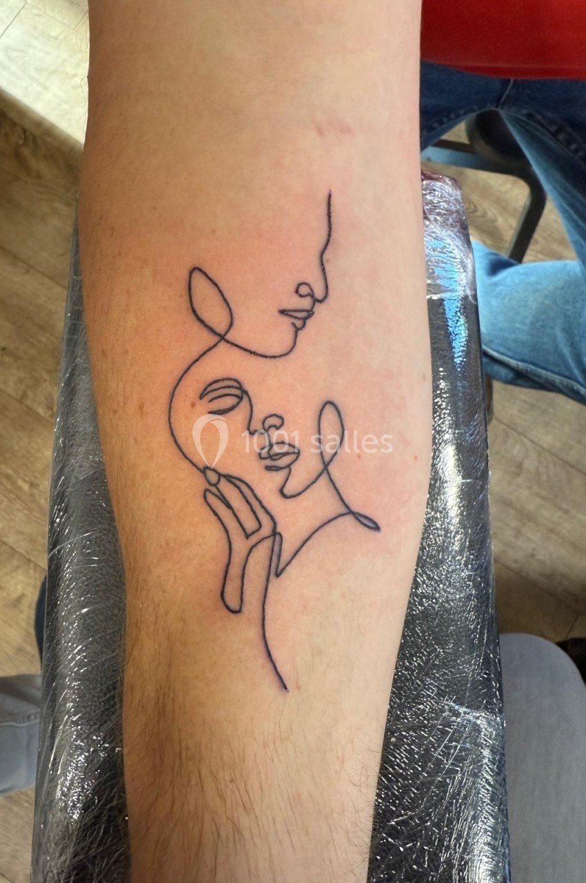 Tatouage minimaliste sur l'avant-bras représentant deux visages entrelacés tracés en ligne continue.