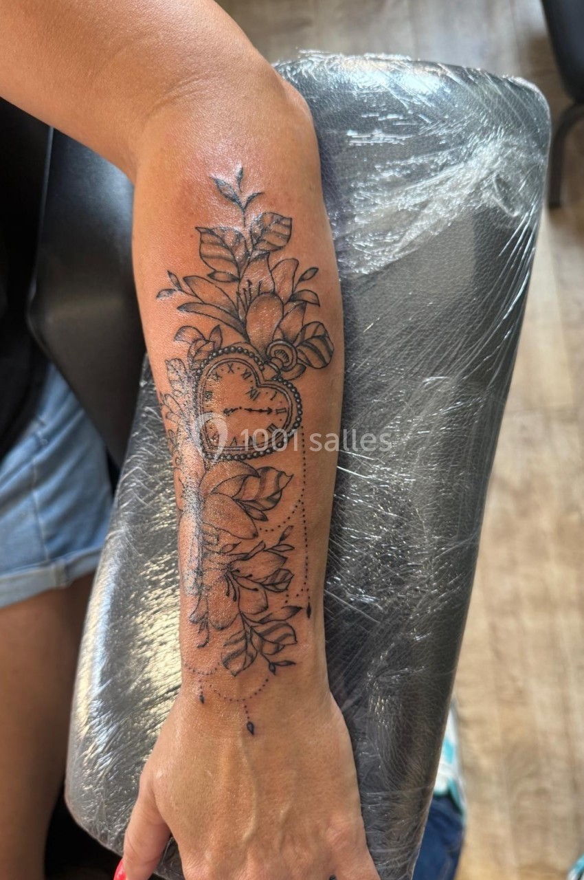 Tatouage détaillé sur l'avant-bras représentant un cœur en forme de montre entouré de fleurs et de feuilles.