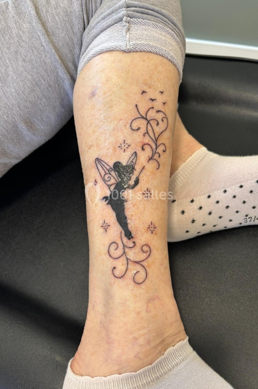 Tatouage d'une fée noire avec des motifs floraux et étoiles, situé sur la jambe d'une personne assise.