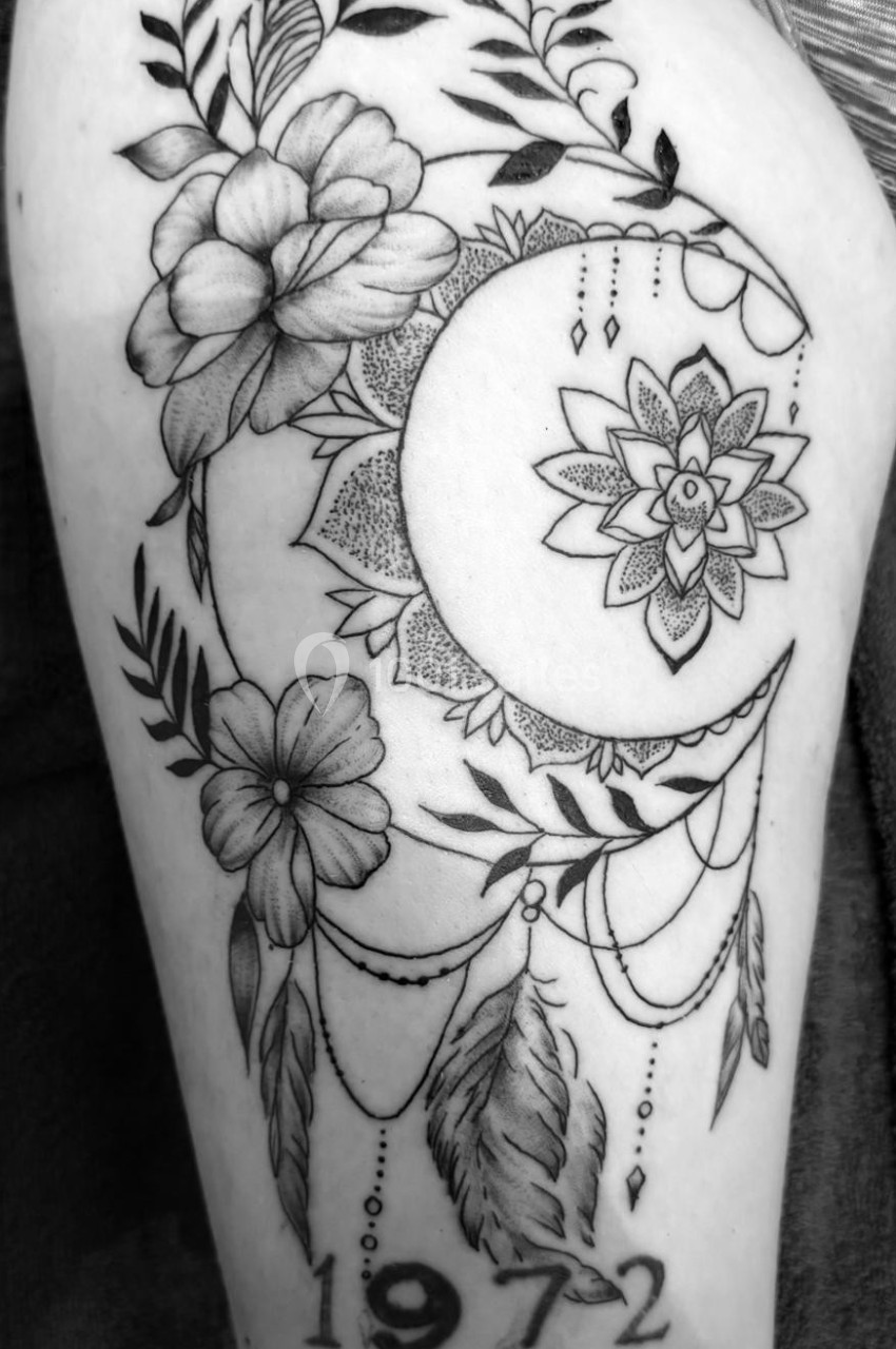 Tatouage représentant une lune ornée de motifs floraux, de plumes suspendues et du chiffre ’1972’.