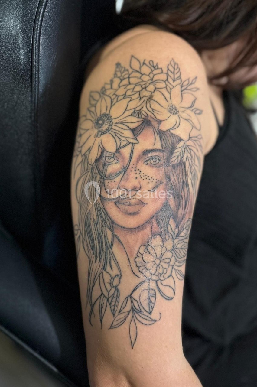 Tatouage sur le bras représentant un visage féminin entouré de fleurs et de feuilles, en noir et gris.