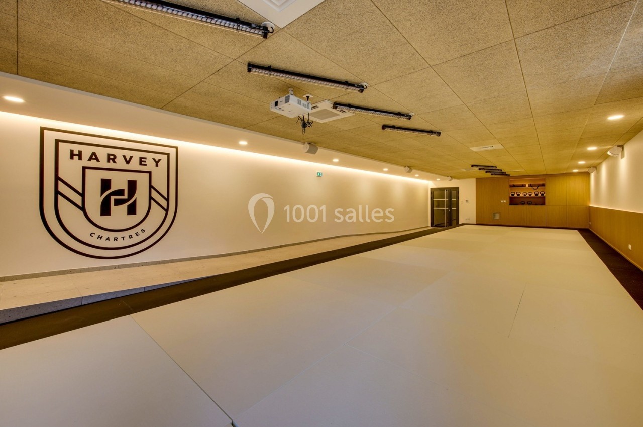 Salle d'entraînement spacieuse avec tatamis au sol, éclairage chaleureux et logo mural ’Harvey Chartres’.