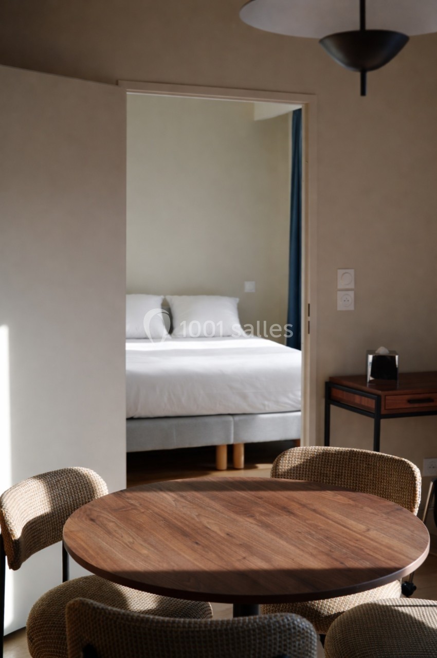 Vue d'une chambre avec un lit blanc, visible à travers une porte ouverte depuis une salle à manger avec table en bois.