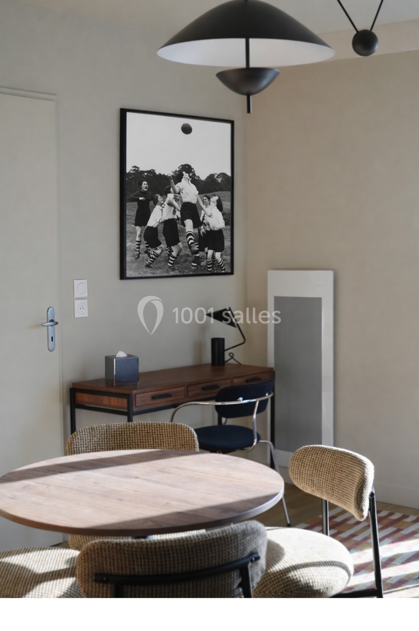 Salle à manger moderne avec table ronde en bois, chaises tissées, bureau en arrière-plan et photo en noir et blanc au mur.