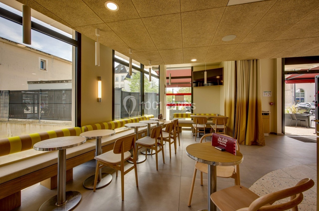 Salle lumineuse avec tables et chaises en bois, grandes fenêtres donnant sur l'extérieur et rideaux jaunes.