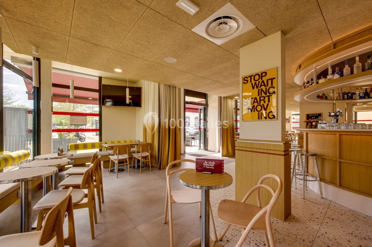 Intérieur d'un café moderne avec tables, chaises en bois, bar à droite et grandes fenêtres laissant entrer la lumière.
