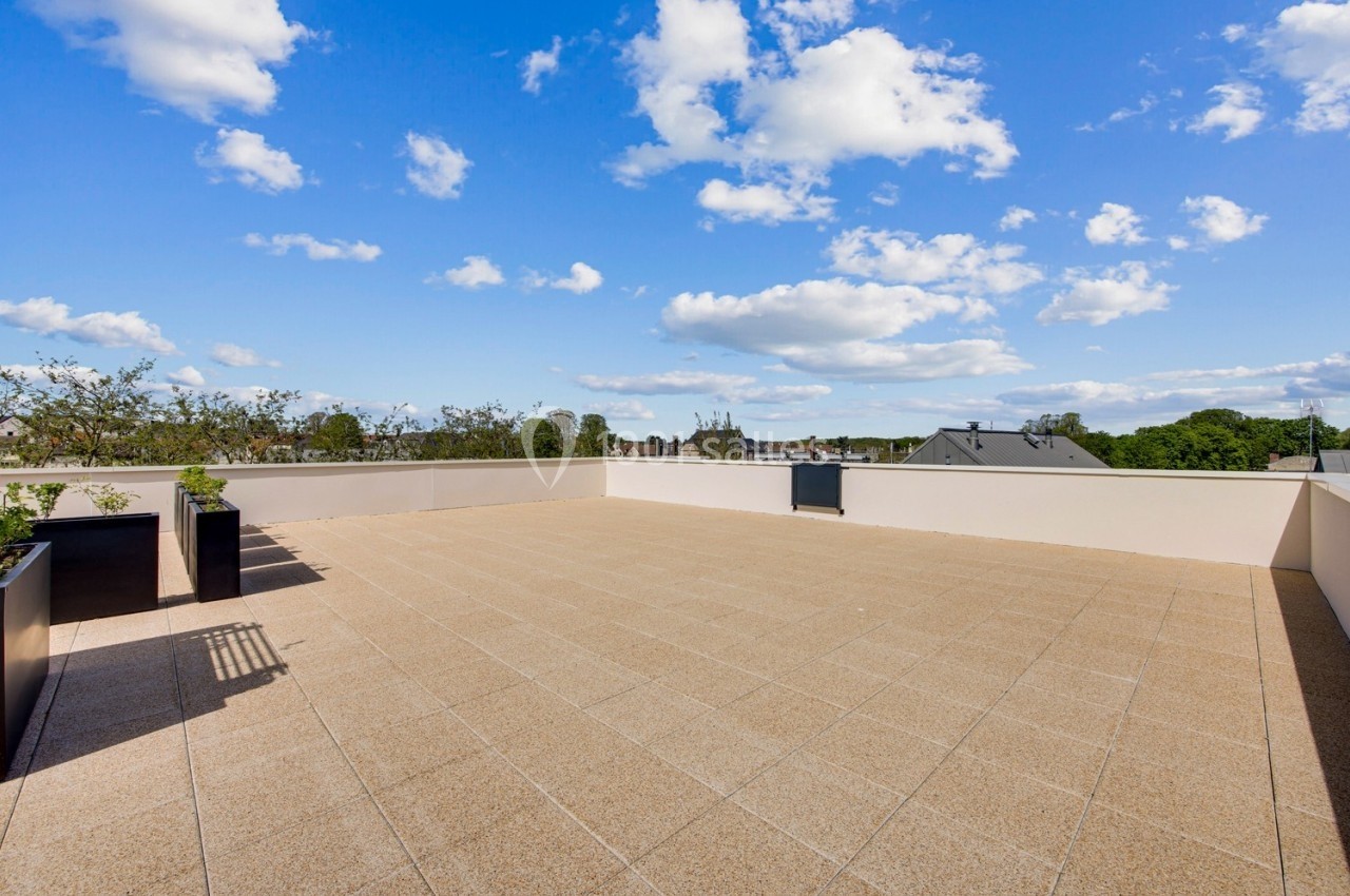 Terrasse spacieuse avec sol en dalles beige, bordée de jardinières, sous un ciel bleu parsemé de nuages.
