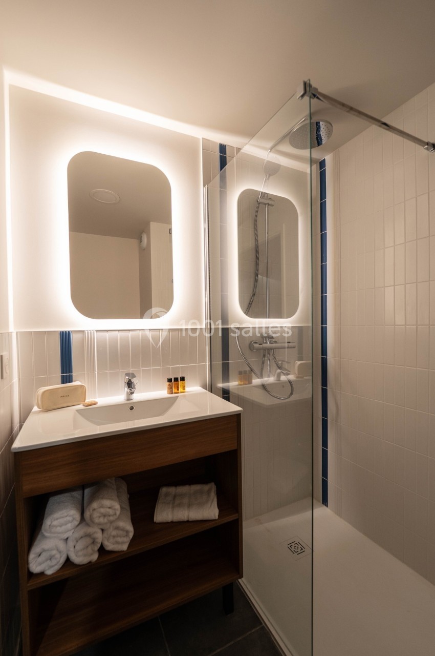 Salle de bain moderne avec douche à l'italienne, meuble en bois avec lavabo, miroir éclairé et serviettes pliées.