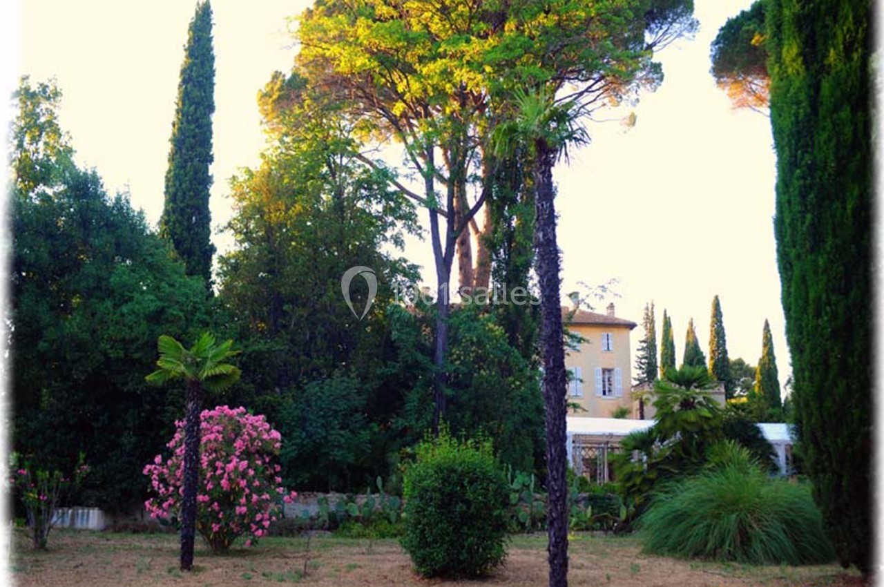 Jardin méditerranéen avec palmiers, cyprès, buissons fleuris et un bâtiment en arrière-plan sous une lumière douce.