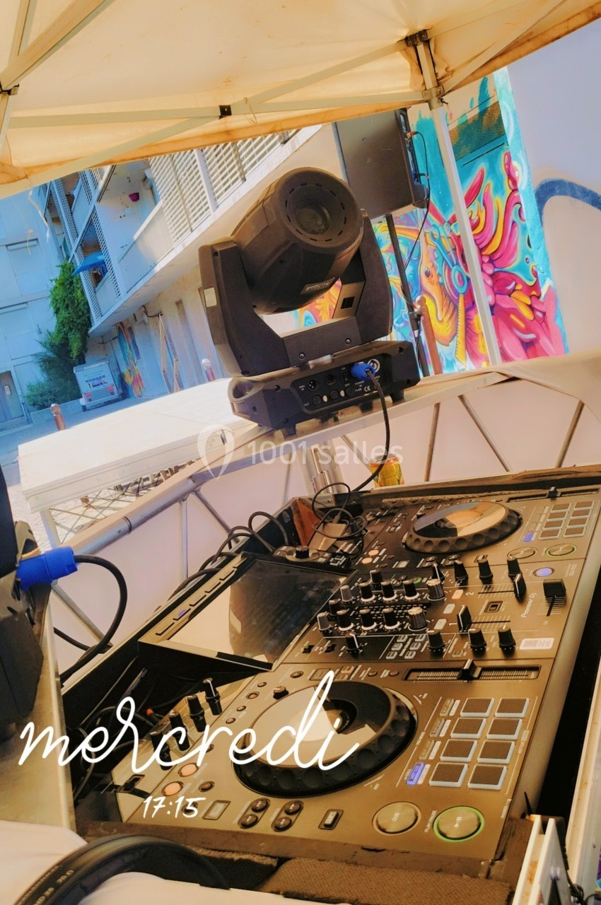 Table de mixage avec platines et projecteur lumineux sous un chapiteau, dans un environnement urbain coloré.