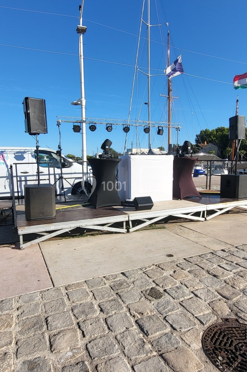 Scène extérieure vide avec équipement audio sur un quai pavé, voilier et ciel bleu en arrière-plan.